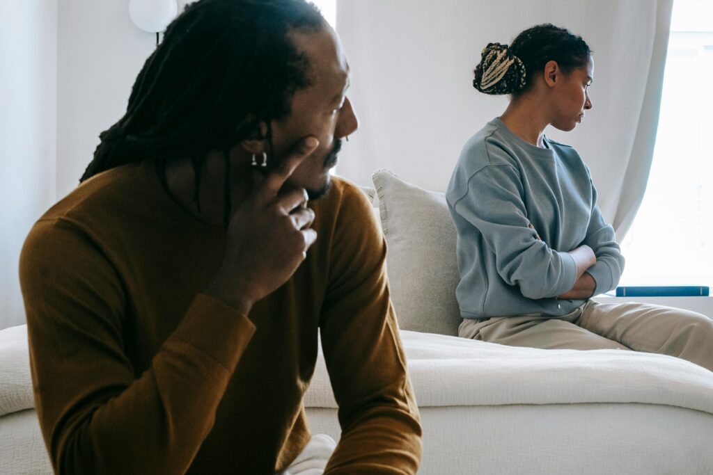 A couple sits on opposite ends of a couch facing away from each other, the man with his head in his hand and the woman with her arms crossed.