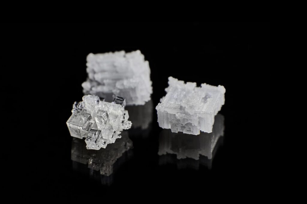 Three translucent white salt crystals with cubic structures sitting on a black reflective surface.