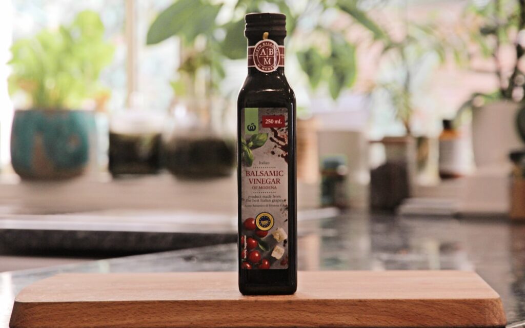 Tall dark bottle of balsamic vinegar standing on a wooden cutting board with houseplants visible in the soft-focus background.
