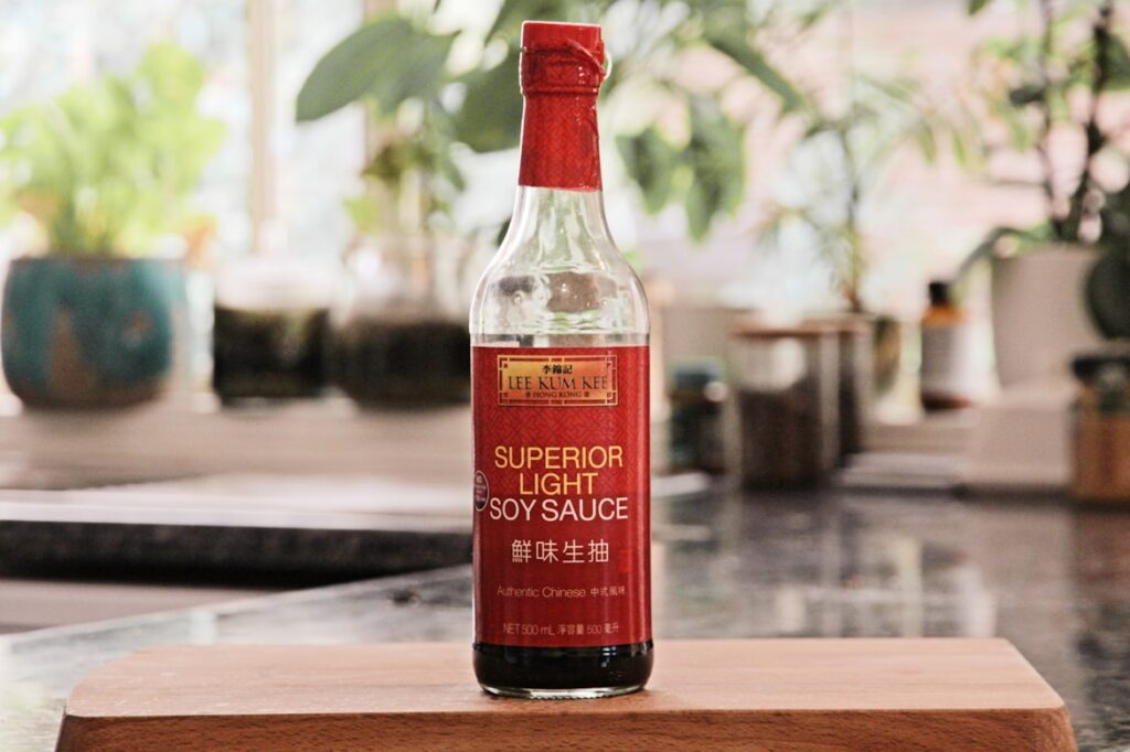 Glass bottle of Lee Kum Kee Superior Light Soy Sauce with red cap and label on a wooden surface, with greenery in the blurred background.