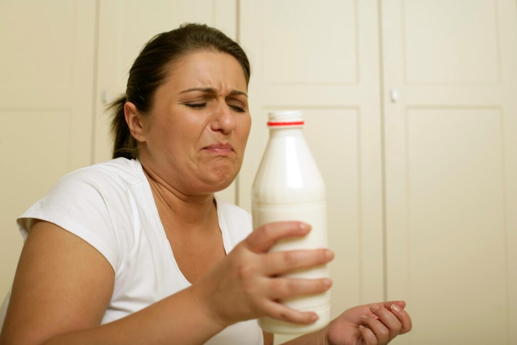 Woman in a white shirt with a disgusted expression on her face while holding a white plastic milk bottle at arm's length.