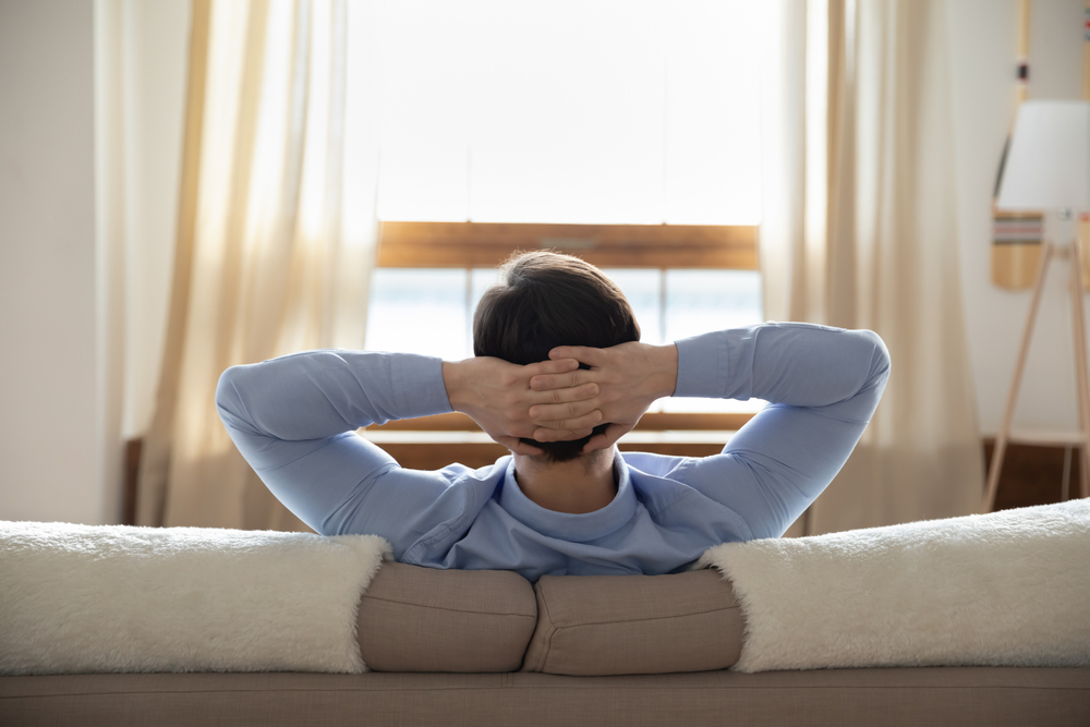 Long lasting plans. Rear view of successful millennial male businessman resting at home sitting on sofa with hands behind head looking at window creating new ideas dreaming of future opportunities