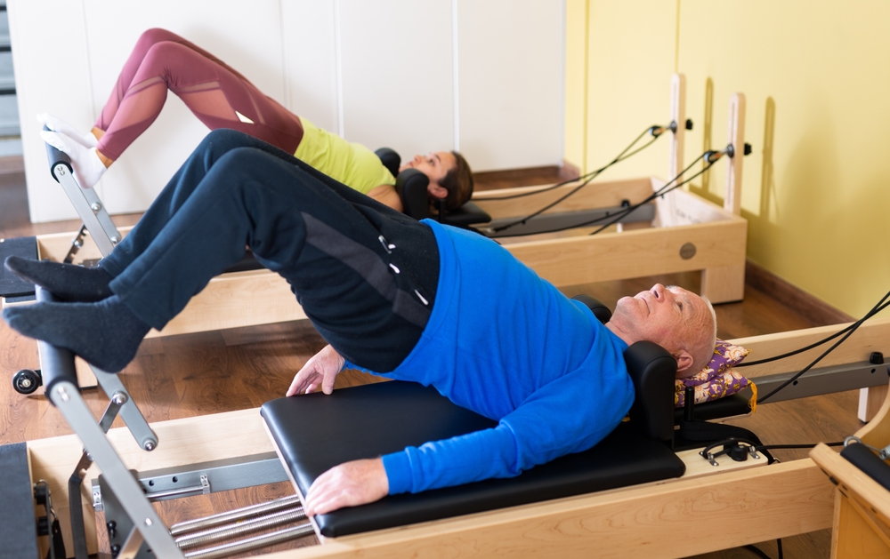 Older man doing exercises of remedial gymnastic on Pilates reformer in rehab clinic. Physical medicine concept ..