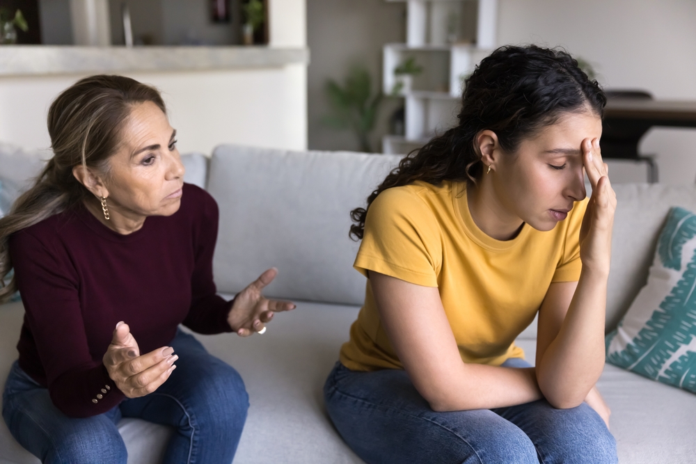 Tense moment between mum and daughter, family conflicts, misunderstanding, toxic relations. Elderly 60s woman scolding, screaming at grown up 20s daughter, fighting, quarrelling at home seated on sofa