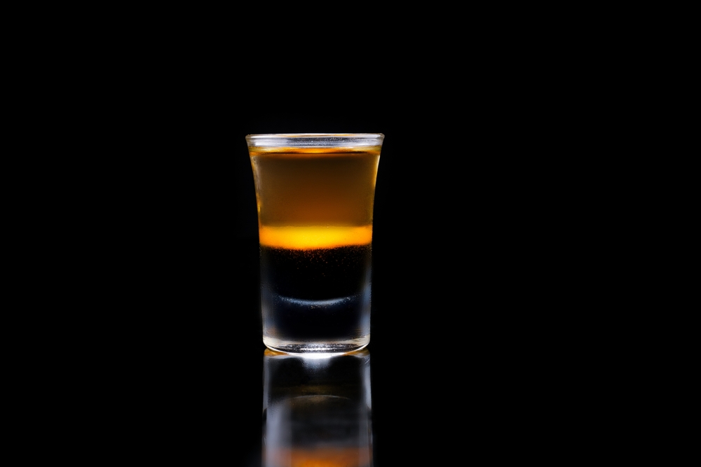 Close-up of a flaming layered cocktail shot in a glass with blue fire on top, isolated on black background with reflection.