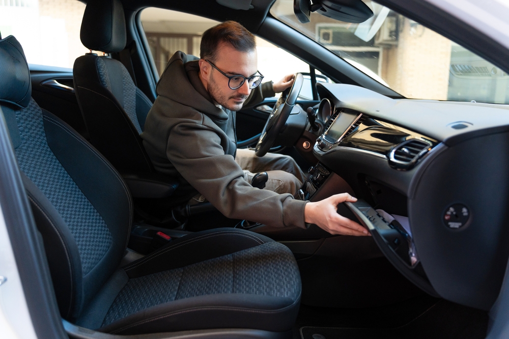 Young man wearing glasses and a hoodie, searching inside the car's open glove compartment, looking for essential items