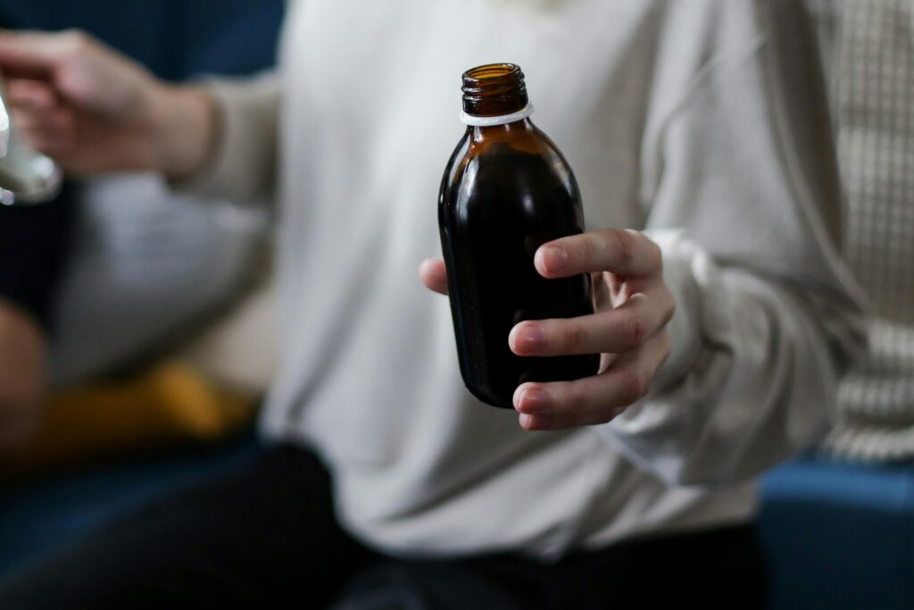A person in a cream sweater holds an uncapped amber glass medicine bottle, the dark liquid visible through the glass. A spoon is visible in the soft-focus background.