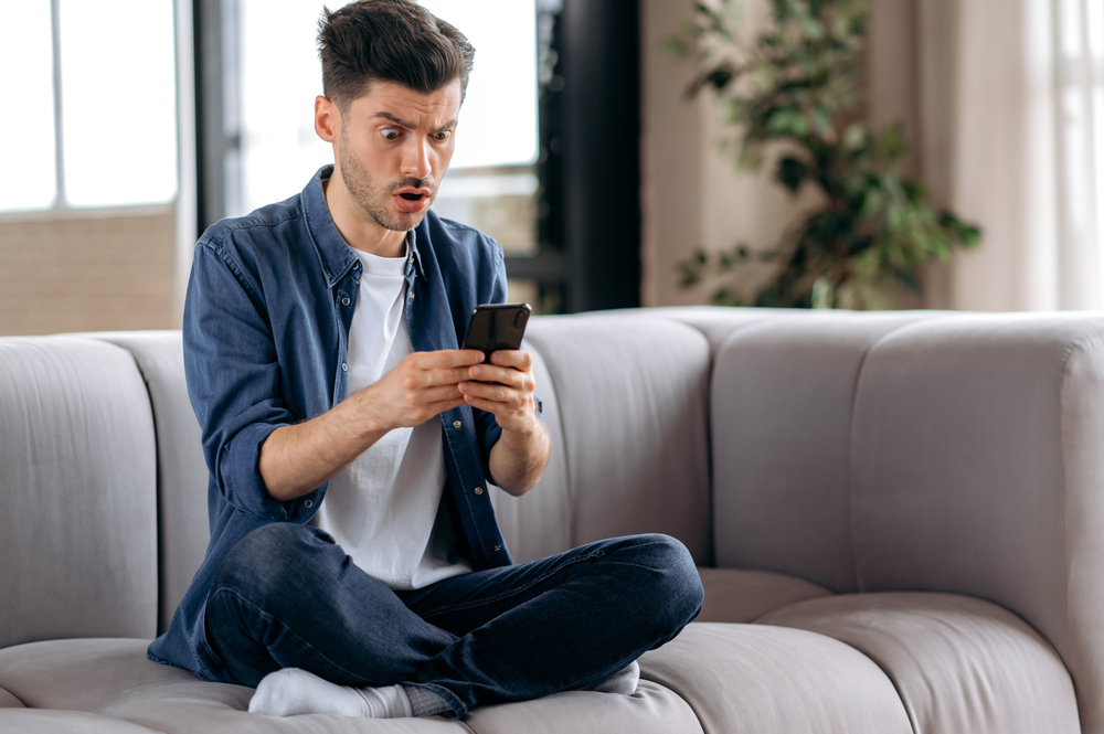 Shocked confused young caucasian man, in casual wear, sitting on sofa in lotus position, uses smart phone, receiving an unexpected message, texting with friends, looking at screen in surprise