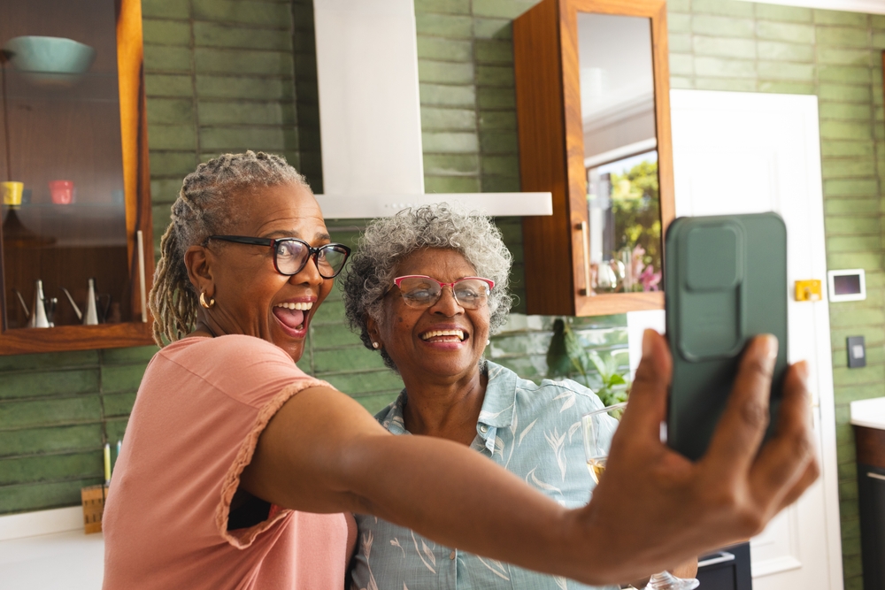 Senior African American woman and senior biracial woman are taking a selfie with a phone. Both are smiling joyfully, capturing a moment of friendship in a cozy kitchen setting.