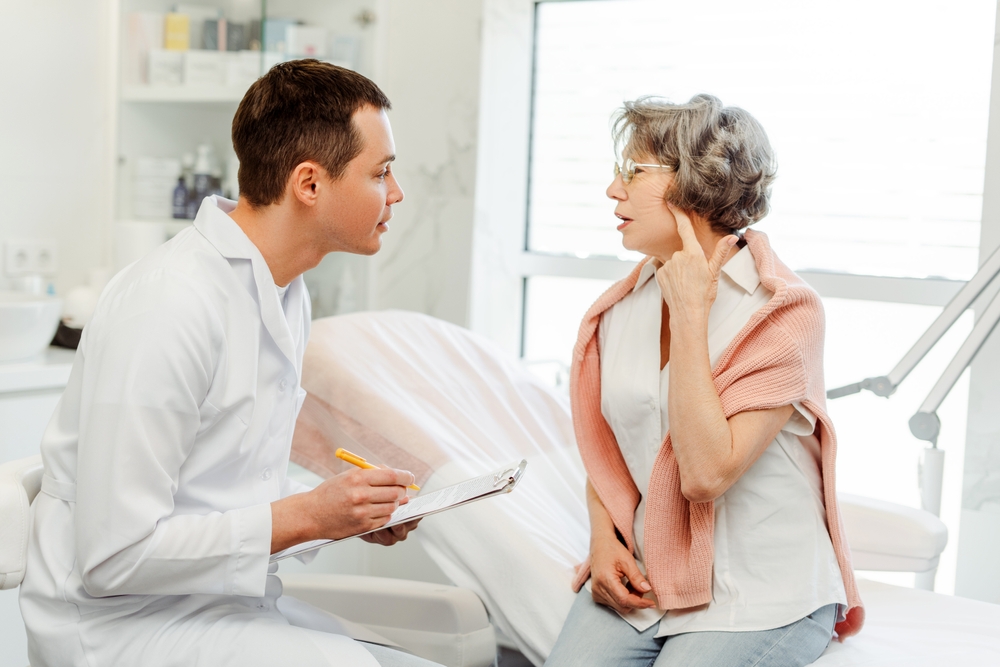 Attractive older gray haired woman talking with man, cosmetologist doctor, pointing finger at face, facelift, sitting in modern clinic. Concept of skin care, treatment, anti-wrinkle procedure