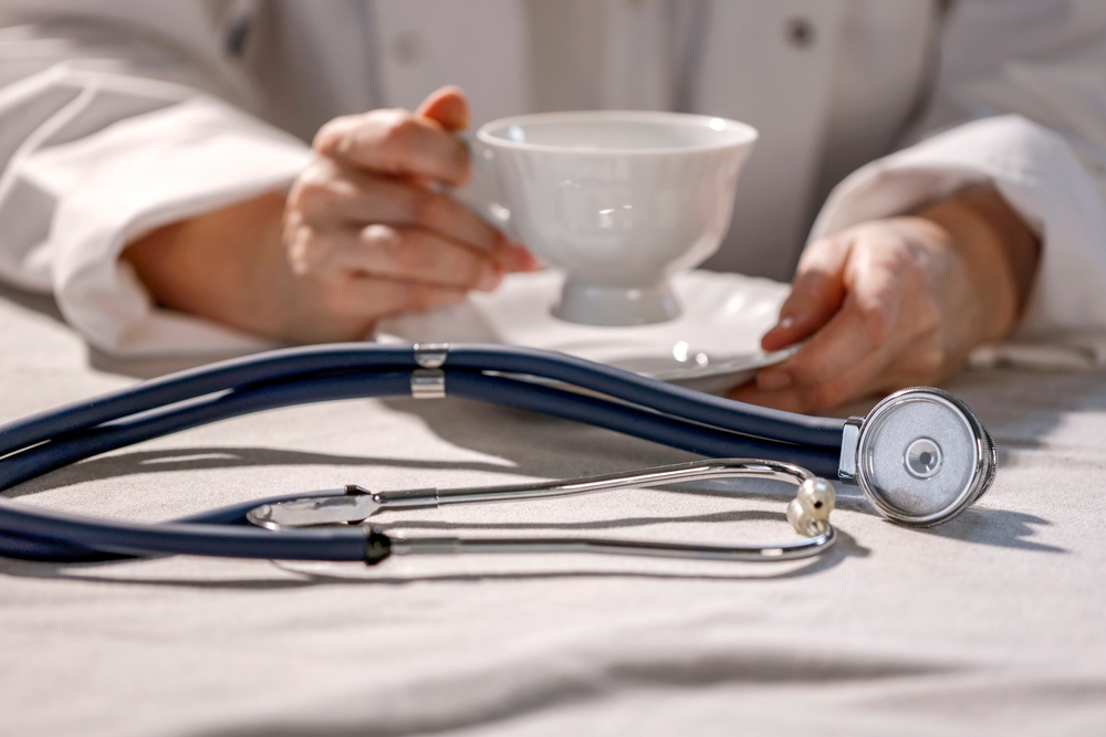 Doctor in white medical coat sitting at table and drinking coffee during break from duty in hospital. Stethoscope foregraound.