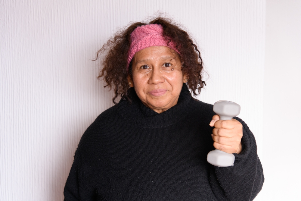 older hispanic latina brunette woman with curly hair and sportswear exercising arms and shoulders with dumbbells at home