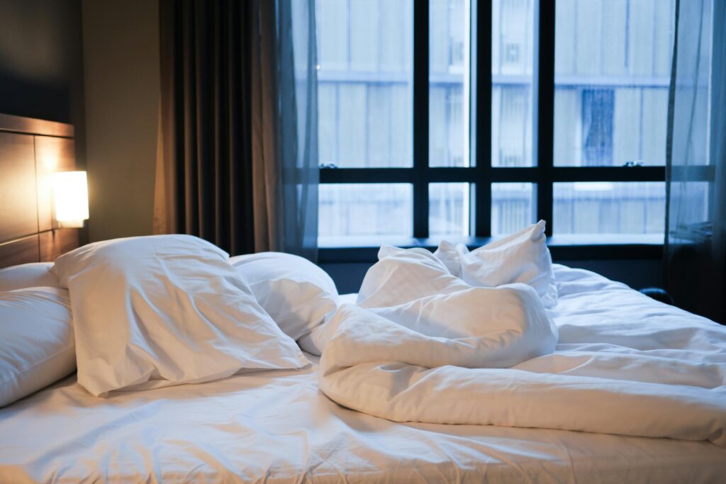 An unmade bed with rumpled white linens and pillows beside a large window.