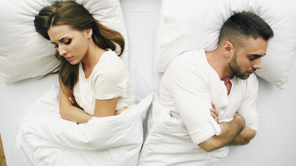 Couple lying back-to-back in bed under white sheets, both looking distant and unhappy.