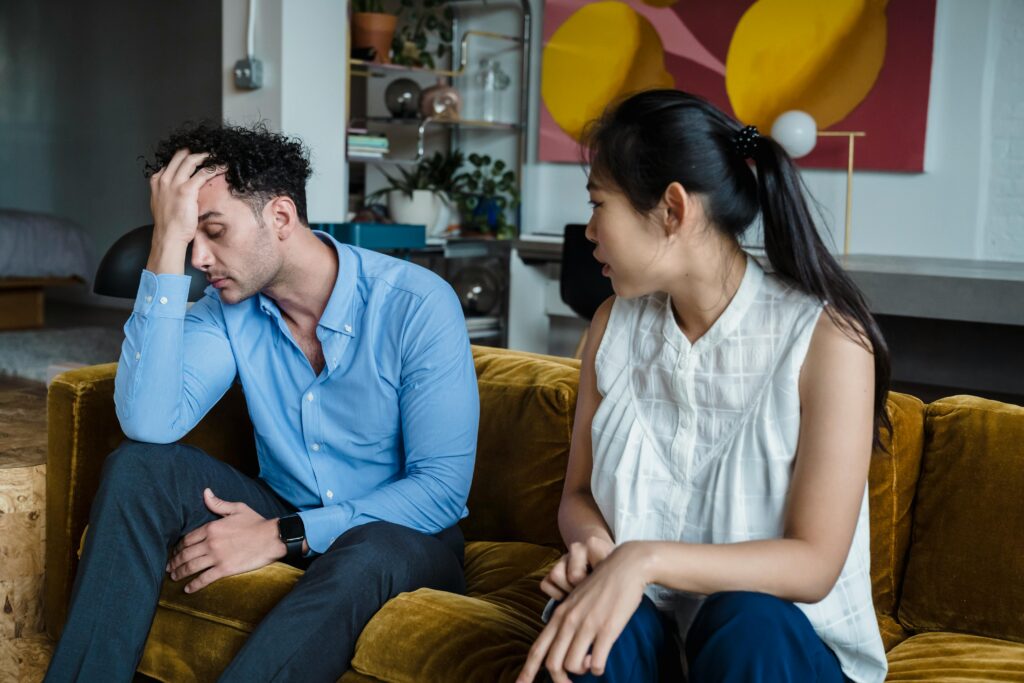 Couple on a yellow couch, the man holding his forehead in distress while the woman watches with concern.