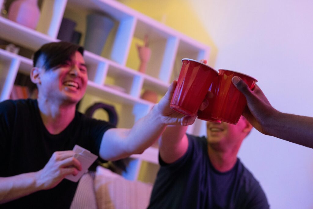 Two men laughing and toasting with red plastic cups in a warmly lit room.