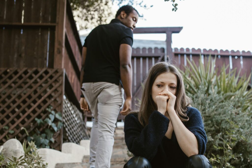 Woman sitting outside with her hands clasped to her face, looking sad as a man walks away behind her.
