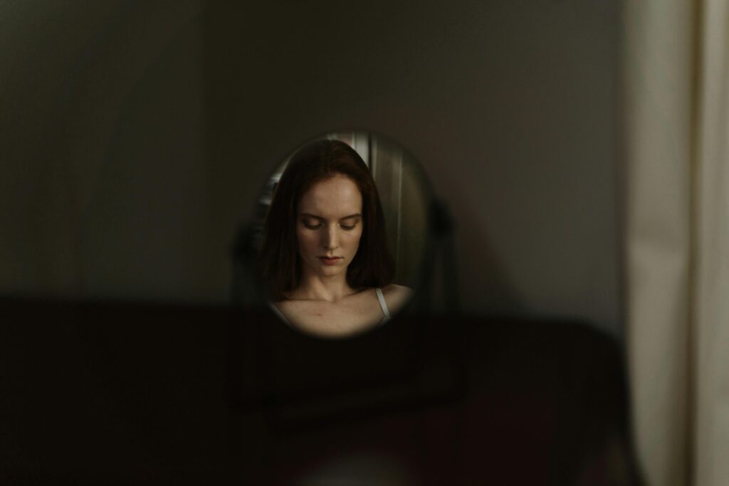 Reflection of a woman with downcast eyes in a small oval mirror, dim warm lighting.