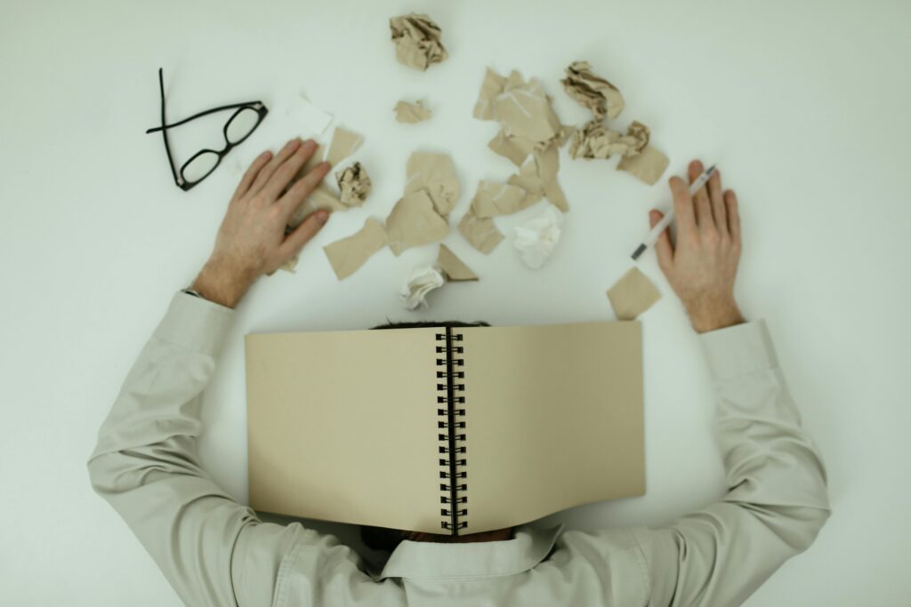 Person slumped behind an open blank notebook on a desk, surrounded by crumpled paper and discarded glasses.