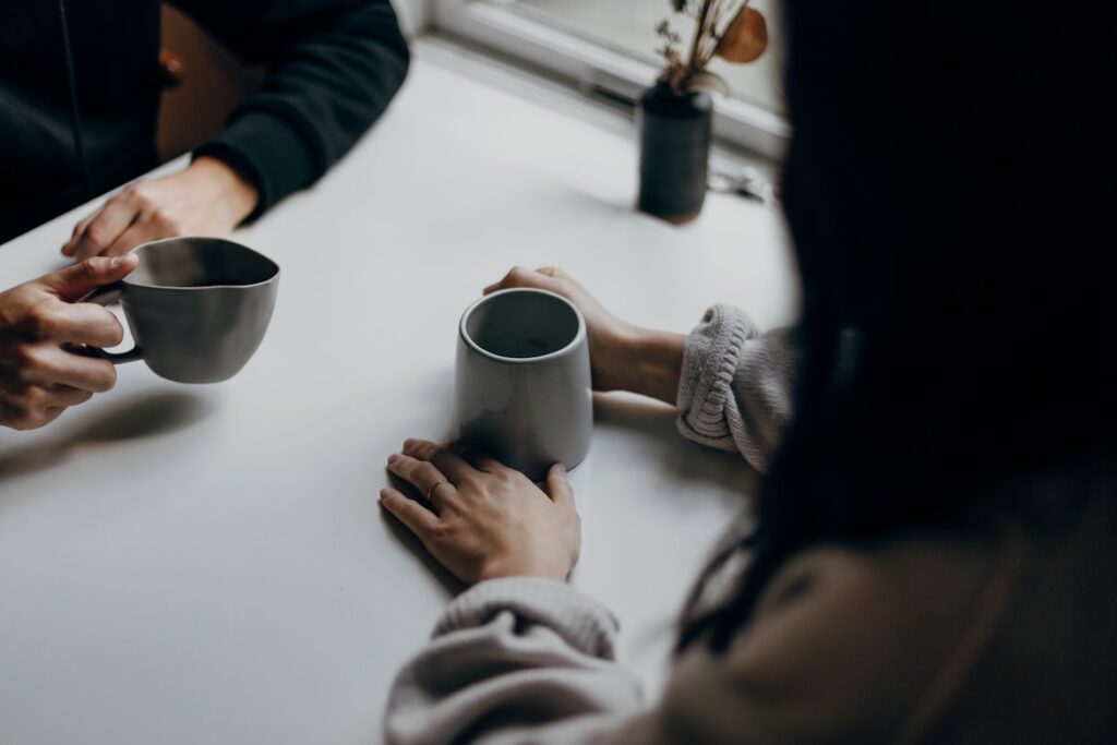 Two people sitting across from each other at a white table, holding coffee mugs mid-conversation.