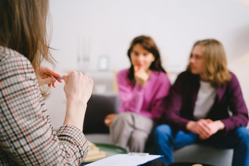 Therapist in the foreground holding a pen while a couple sits across from her, listening.