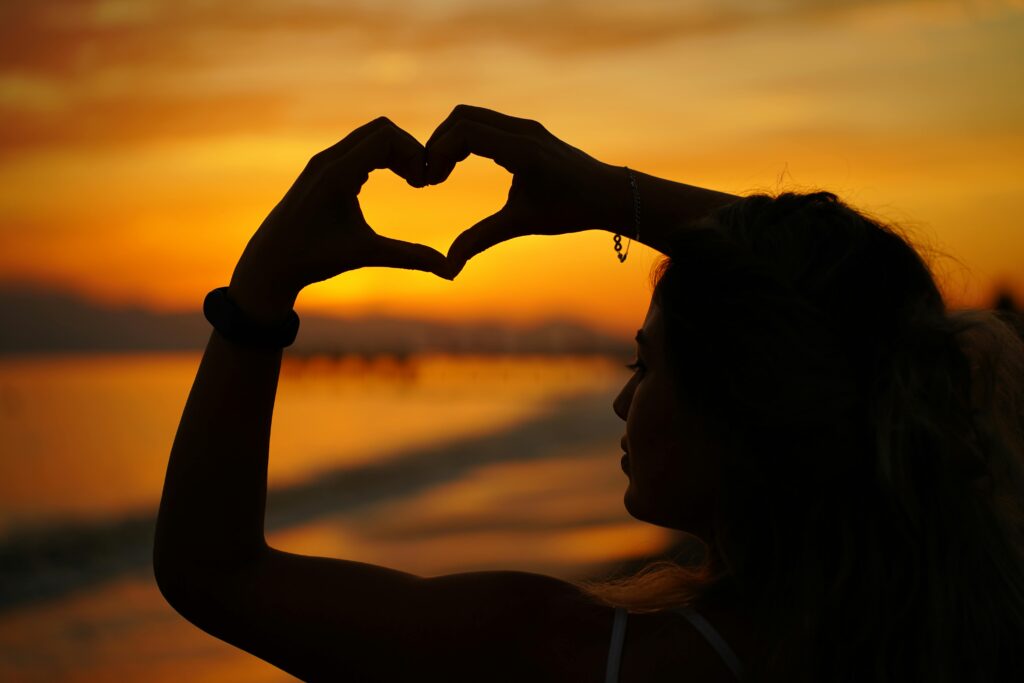 Claude responded: Silhouette of a woman on a beach forming a heart shape with her hands against a glowing orange sunset over the ocean.Silhouette of a woman on a beach forming a heart shape with her hands against a glowing orange sunset over the ocean.