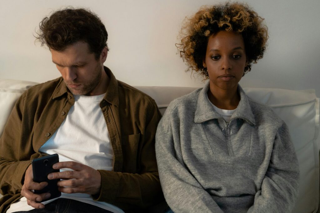 Couple sitting on a couch, the man scrolling his phone while the woman stares ahead with a sad expression.