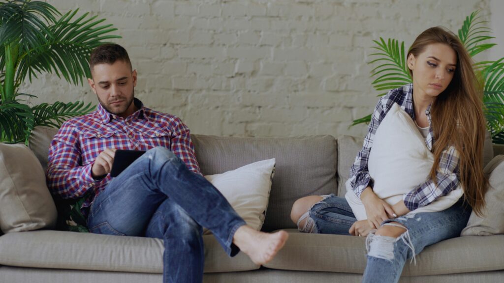 Couple on a sofa, the man absorbed in his tablet while the woman hugs a pillow and looks away.