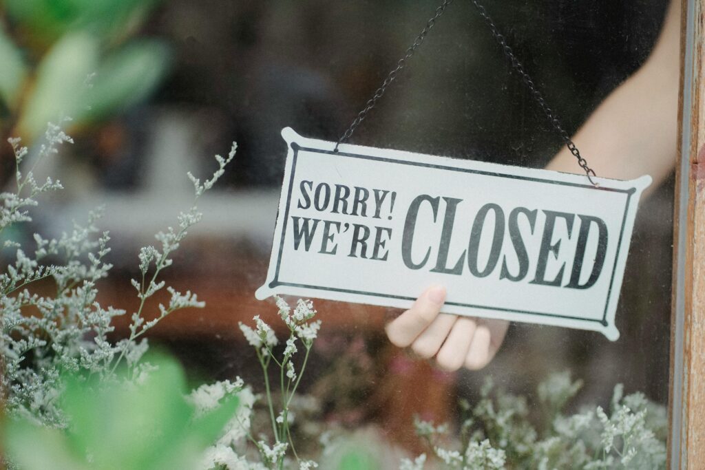 Hand holding a Sorry We're Closed sign in a shop window framed by soft greenery.