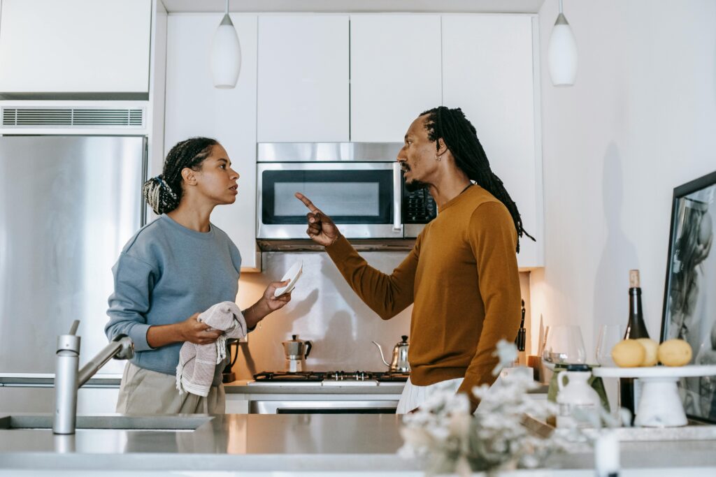 Couple arguing in a kitchen, the man pointing his finger while the woman holds a plate and dishcloth.