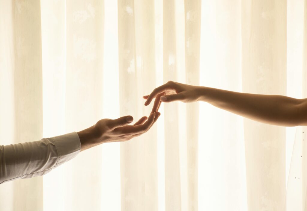 Two hands reaching toward each other against soft sheer curtains, fingertips almost touching.
