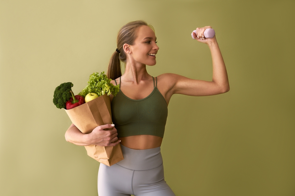 Sporty beautiful woman holding a bag of vegetables and a dumbbell on a green background. The concept of sports and healthy eating.