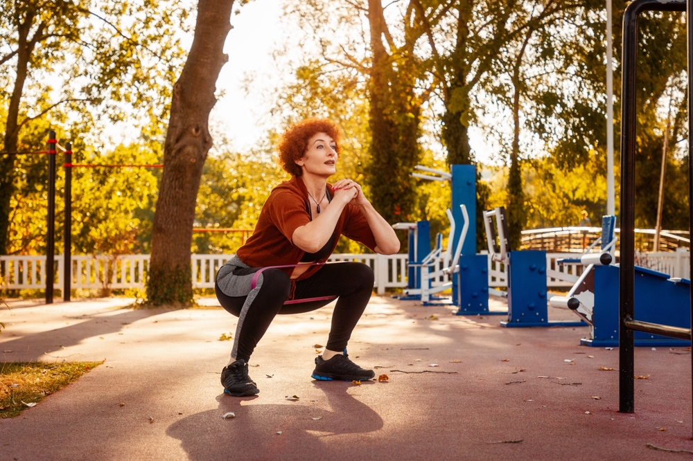 Adult Caucasian woman with makeup squatting with elastic band in sports park. Concept of healthy lifestyle and fitness.