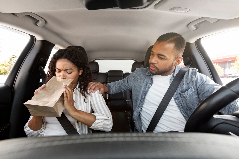 Sick black lady sitting in the front seat of a car covers mouth, breathing in paper bag. Dizzy african american woman suffers from motion sickness during road trip, concerned man husband comfort her