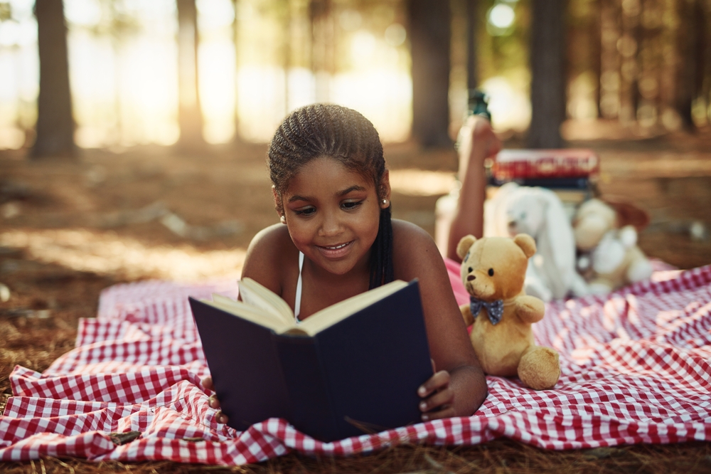 Happy African kid, reading book and relax outdoor for learning, study or education in forest. Picnic blanket, park and girl with story, magic fantasy or fairytale knowledge on holiday with teddy bear