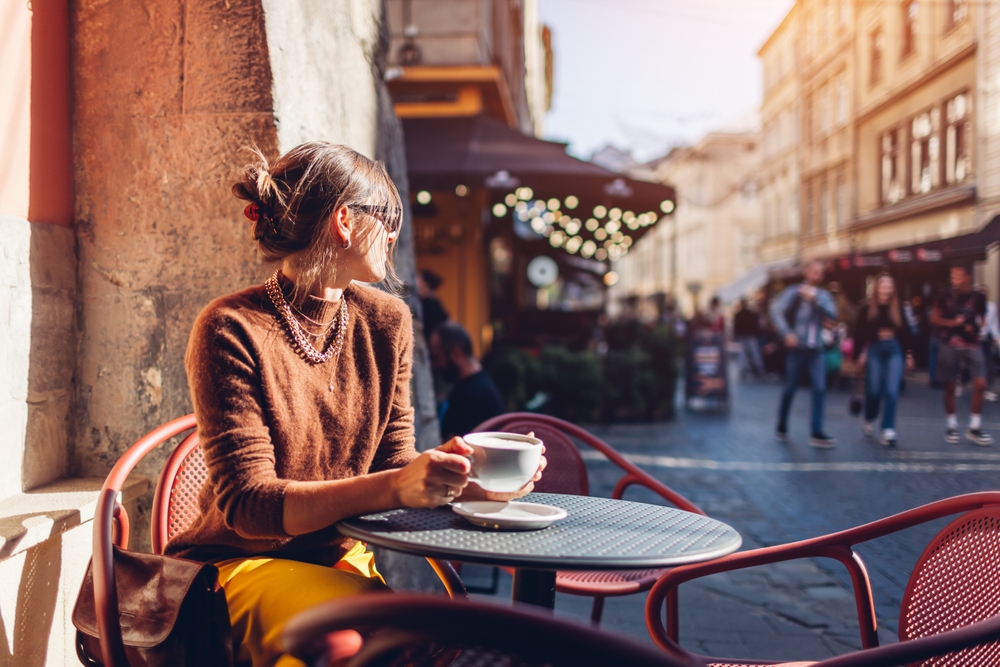 Portrait of stylish woman sitting in cafe outdoor drinking coffee. Woman enjoying architecture of Lviv city on street on autumn day