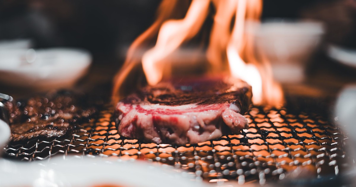 Close-up of a steak being grilled on a charcoal BBQ with visible flames, creating an appetizing view.