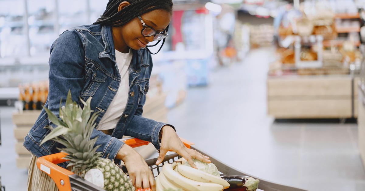A woman shops for fresh fruits and vegetables in a supermarket, pushing a cart filled with groceries.