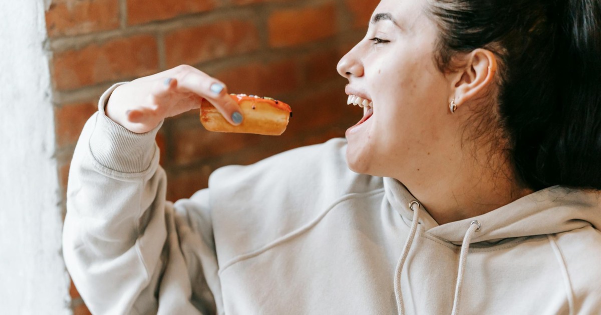 Crop joyful female wearing loose hoodie biting tasty fresh donut in light room