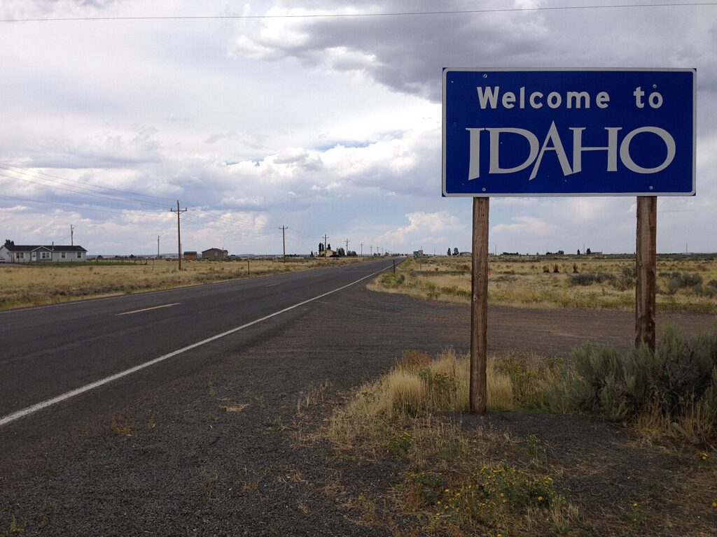 A blue "Welcome to Idaho" highway sign on wooden posts along a flat, arid road stretching into the distance under a cloudy sky.