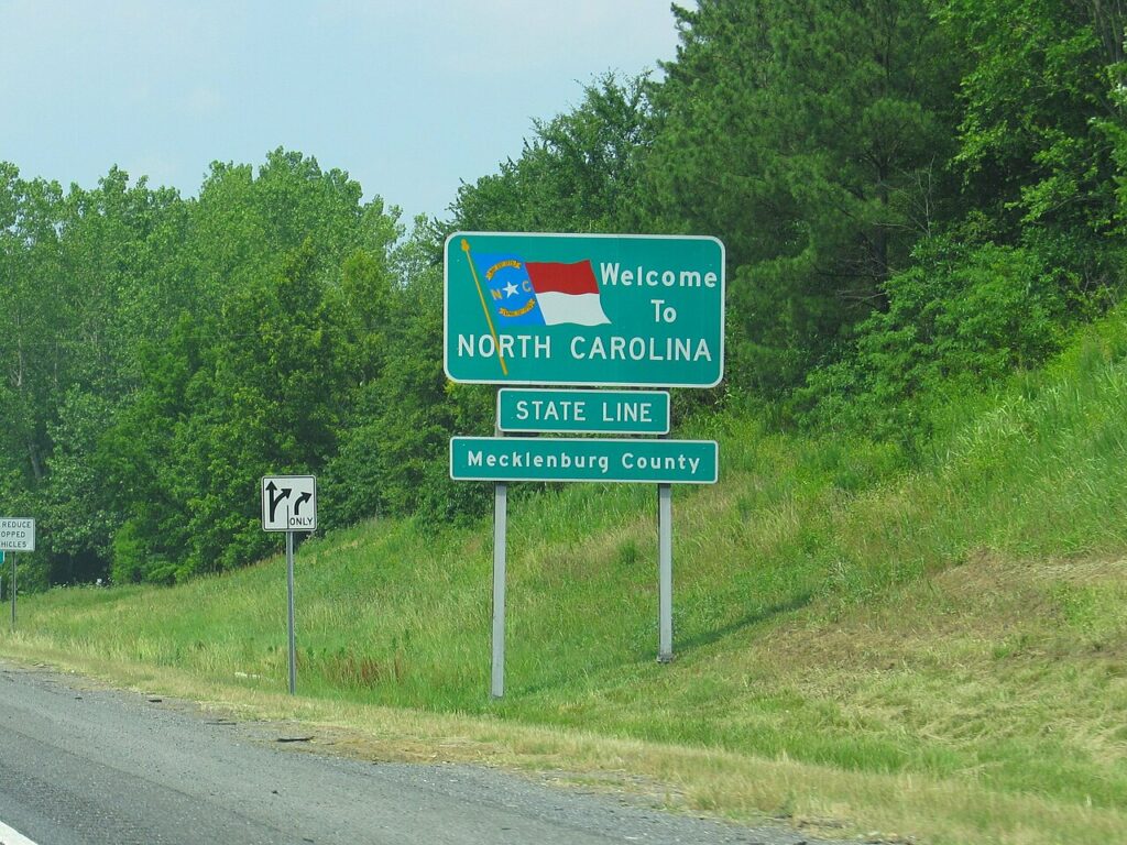 A green "Welcome to North Carolina" highway sign with the state flag, marking the state line at Mecklenburg County along a grassy roadside.