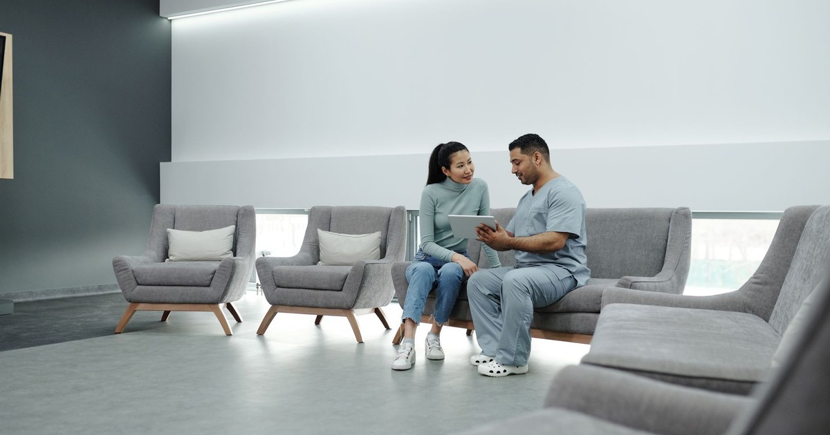 A doctor in scrubs consulting a female patient in a modern clinic waiting room, using a tablet.