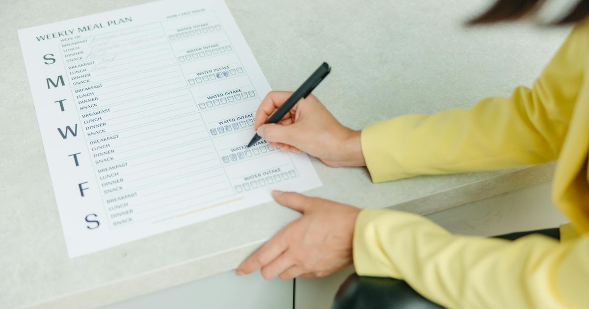 Woman writing on a weekly meal plan checklist indoors, focusing on diet organization.