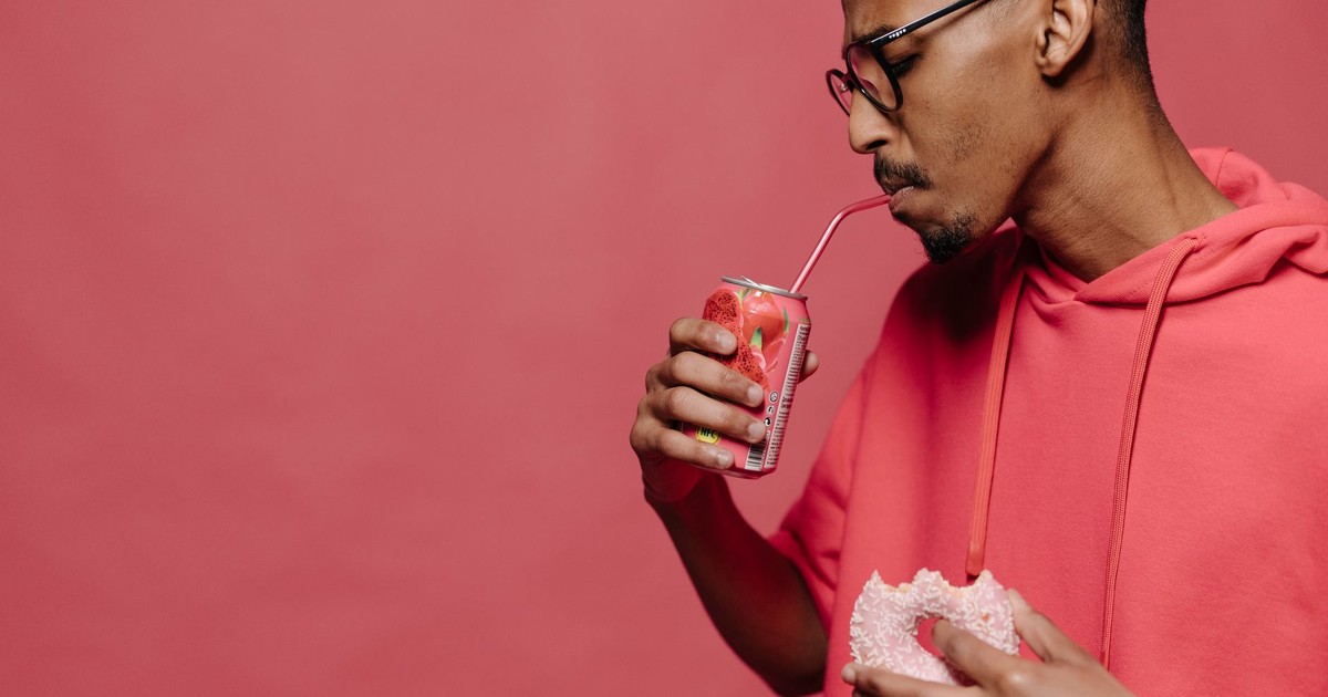 A stylish man drinks soda and holds a donut in a red themed studio setting.
