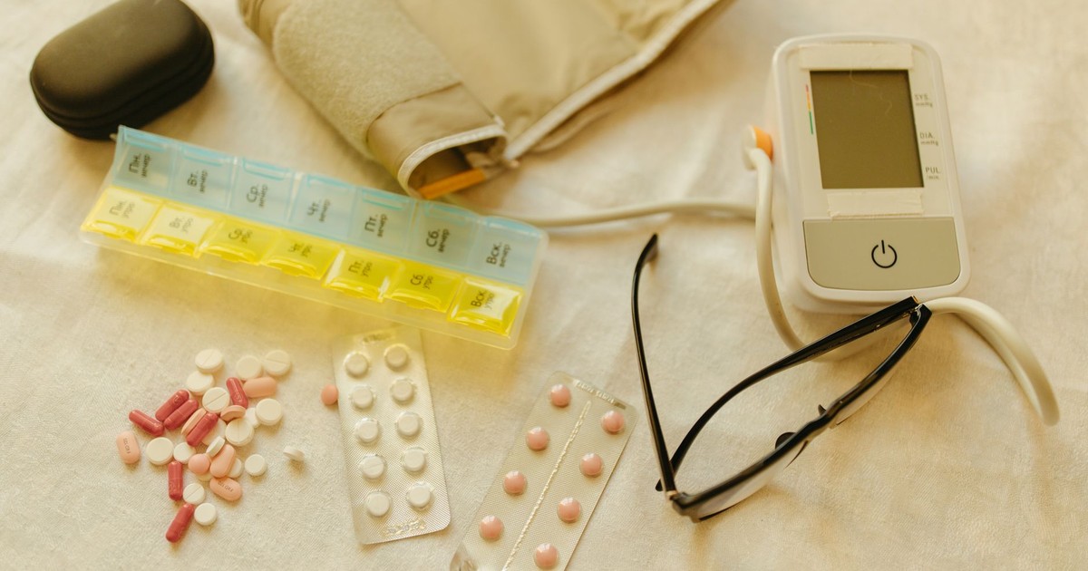 Flat lay image of healthcare essentials including a blood pressure monitor, pills, and eyeglasses on a white surface.