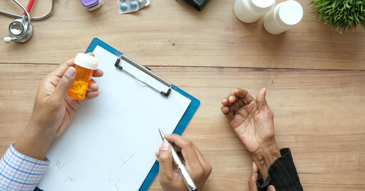 A doctor holds a pill bottle while consulting with an elderly patient. A stethoscope and prescriptions are visible.