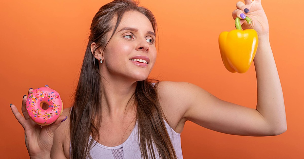 Woman in sports bra juggling choice between a donut and bell pepper on orange background.