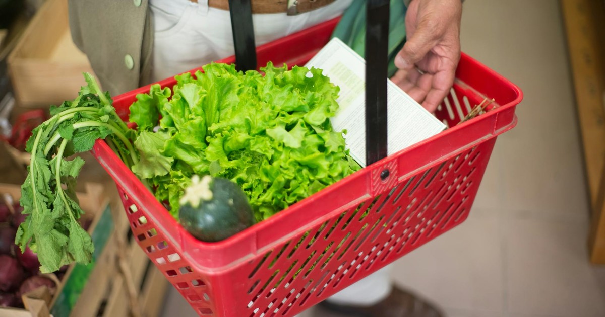 Close-up of a hand carrying a shopping basket filled with fresh greens in a market.