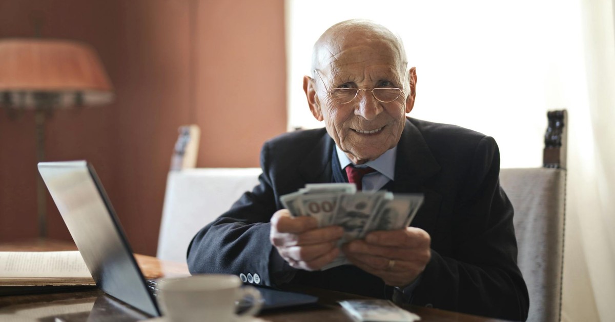 Positive senior businessman in formal suit and eyeglasses holding money in hands while sitting at table near laptop and looking at camera