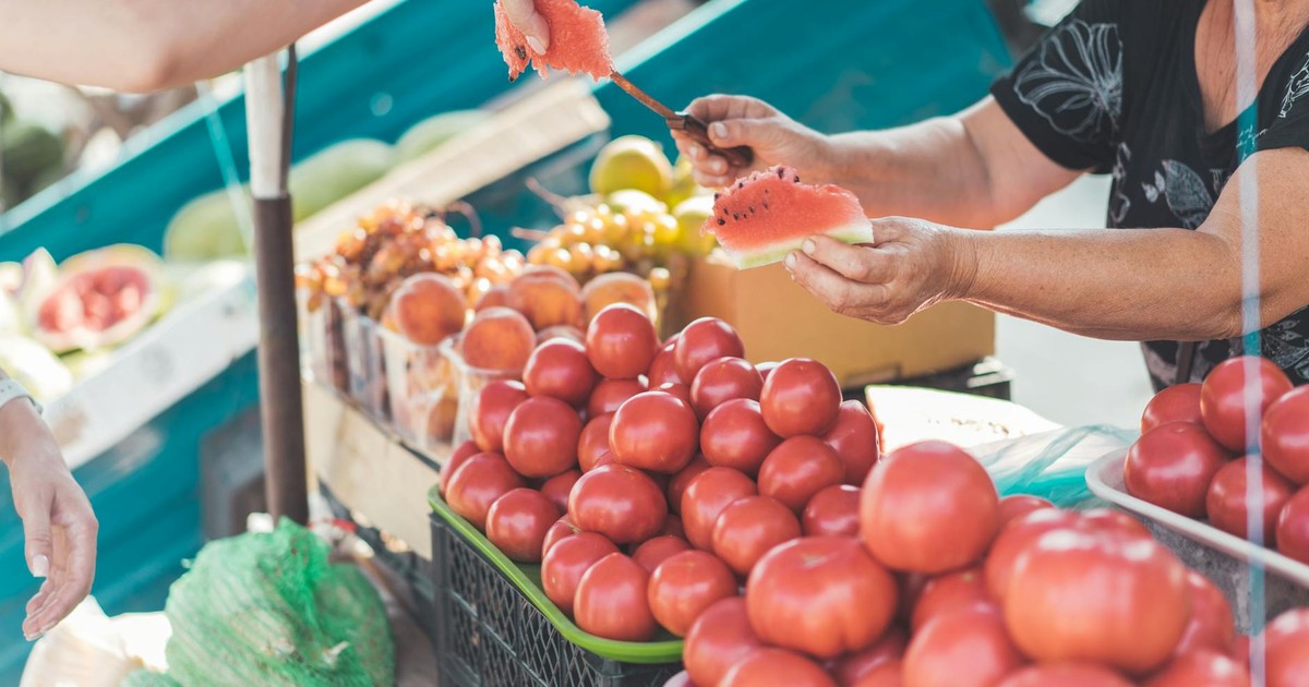 A vendor hands a watermelon slice at a vibrant outdoor market with various fruits.